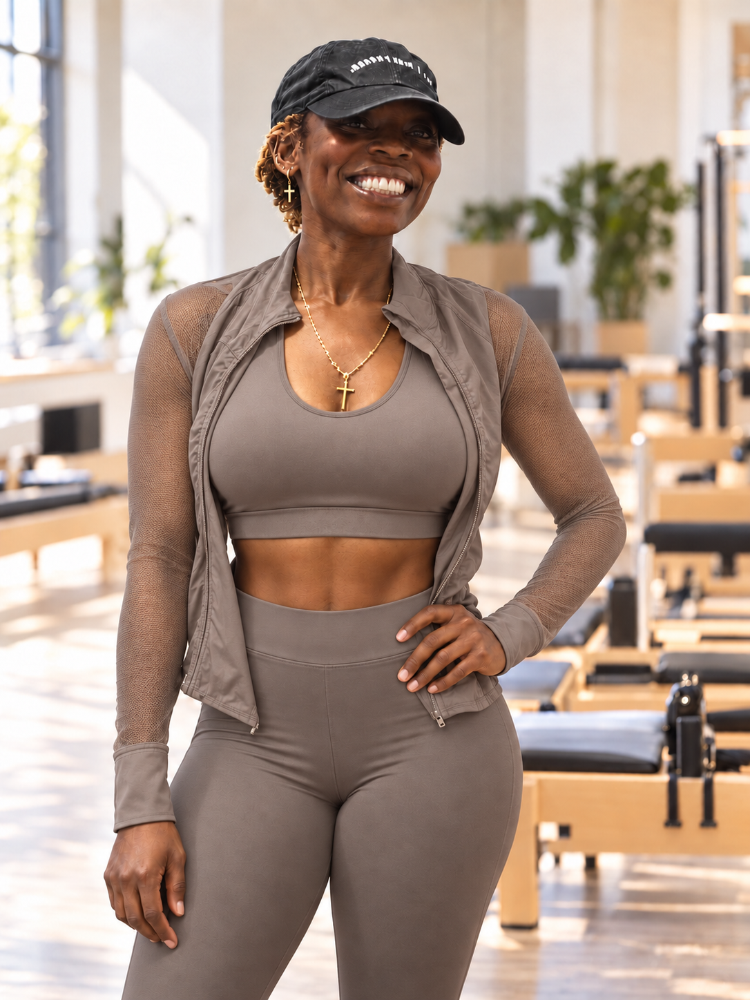 EJ smiling in a Pilates studio, wearing a taupe activewear set and a cap, reformers behind her